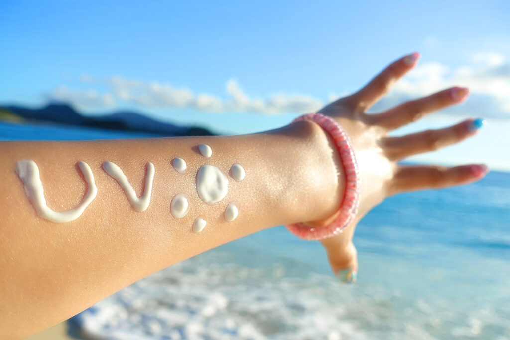 腕に日焼け止めクリームでUVという文字を書き、海を背景に腕を撮影している画像