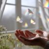 窓辺の光の中で、手のひらから羽のような蝶がそっと飛び立つ様子。