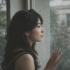 窓辺に立ち、静かに外を眺める女性の横顔。