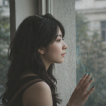 窓辺に立ち、静かに外を眺める女性の横顔。